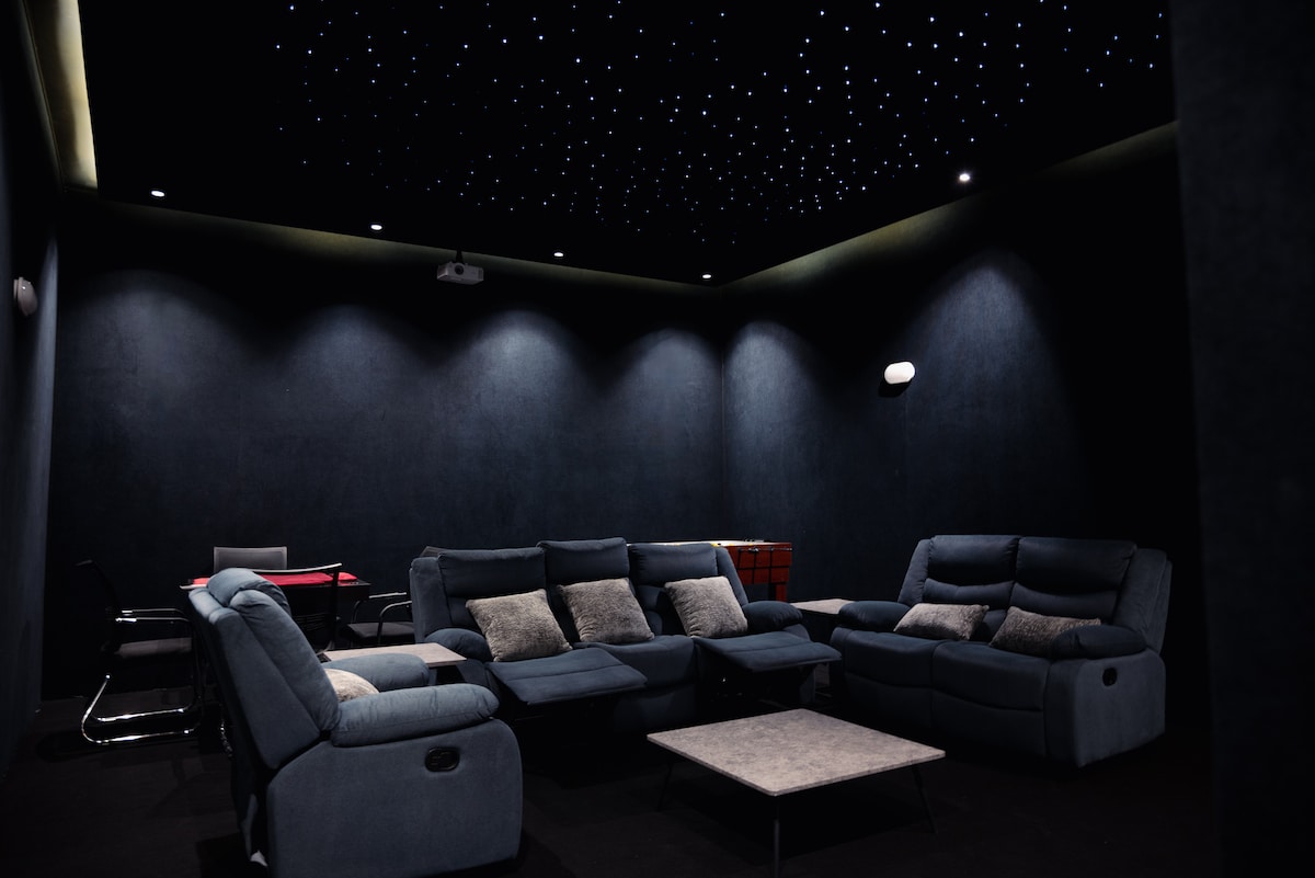 A private cinema room is furnished with comfortable seating arranged around a central coffee table. The ceiling features dazzling star-like lighting, creating a cozy atmosphere. A billiards table is visible in the background, along with some dining chairs set against the dark-toned walls.