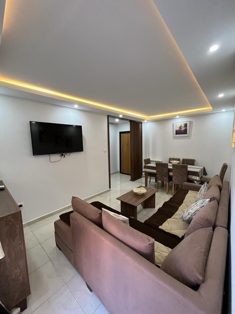 A modern living area features a comfortable sofa positioned facing a wall-mounted television. Soft lighting accents the ceiling, while a dining table is set nearby, surrounded by chairs. Neutral tones and light tiles create a clean, inviting space.