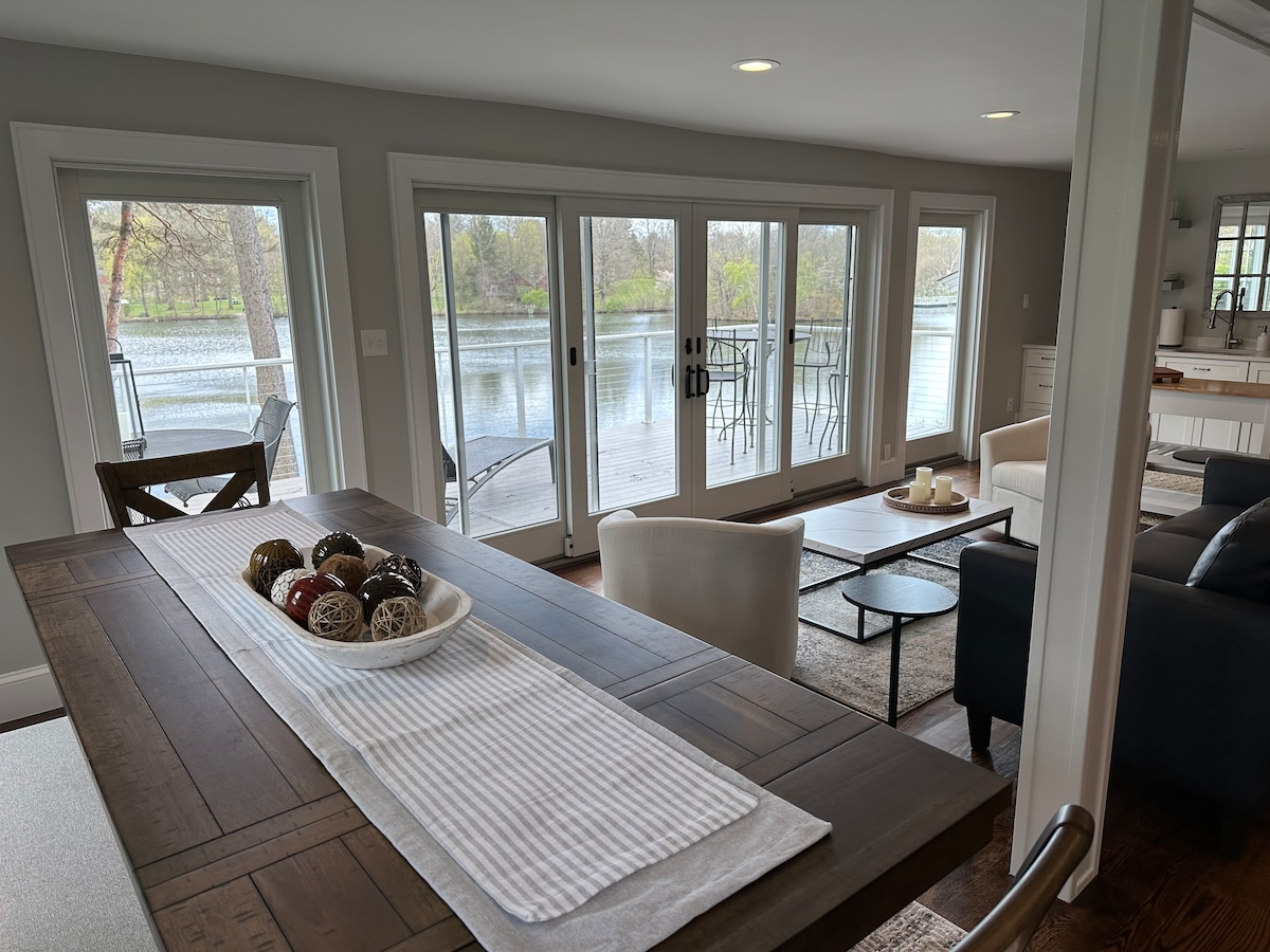A dining table is set with decorative items, positioned neatly under a wide window that offers a view of the lakeside. The room features a mix of seating areas, with a light color palette and natural light reflecting off the water.
