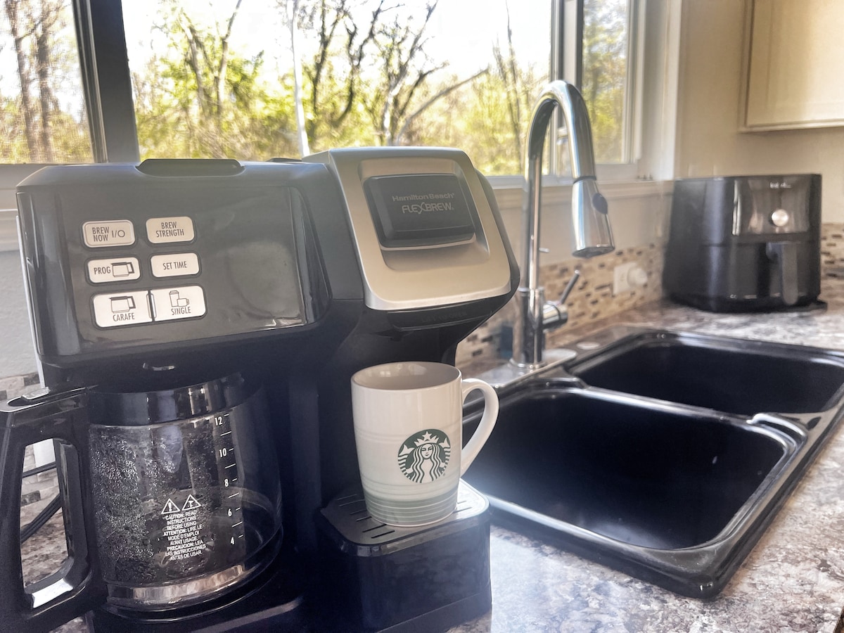 A modern kitchen countertop is featured with a coffee maker alongside a glass carafe and a Starbucks mug. In the background, a double sink is visible, accompanied by a sleek black appliance, enhancing the functional design of the space.