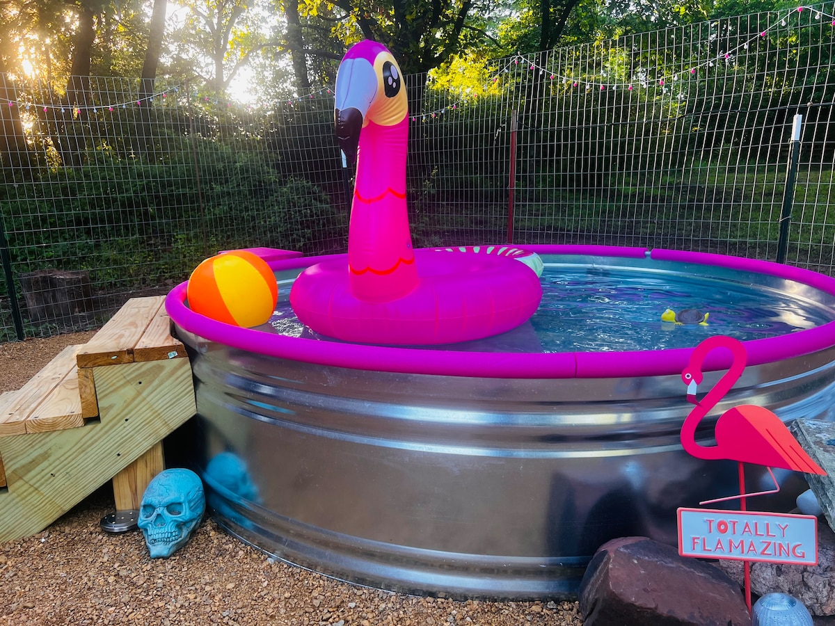 A circular metal stock tank pool is surrounded by a wooden deck. A large inflatable flamingo rests on the water's surface, alongside a colorful beach ball. The pool area is enclosed by a fence, with trees and greenery visible in the background.