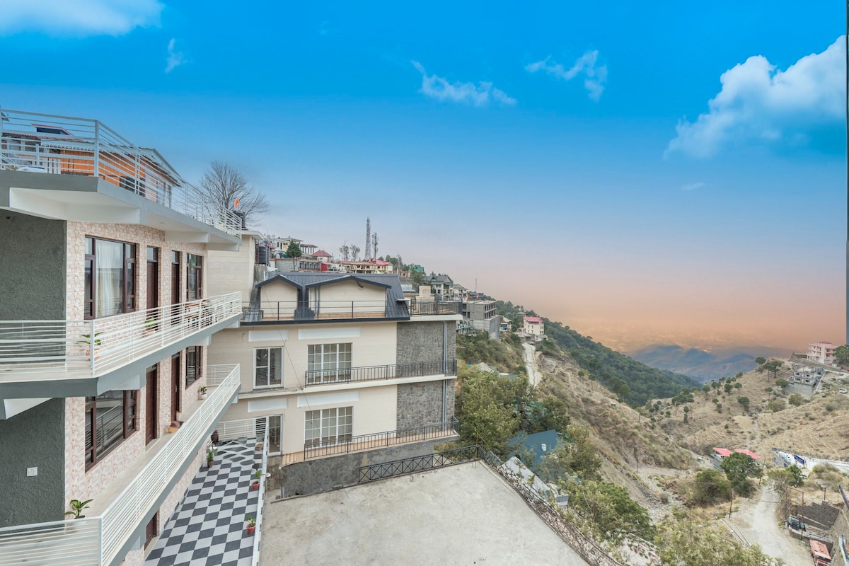 A multi-level building is shown with a modern facade, complemented by a spacious terrace. A view of rolling hills and a clear sky is visible in the background, while a checkerboard pattern covers the patio area beneath.