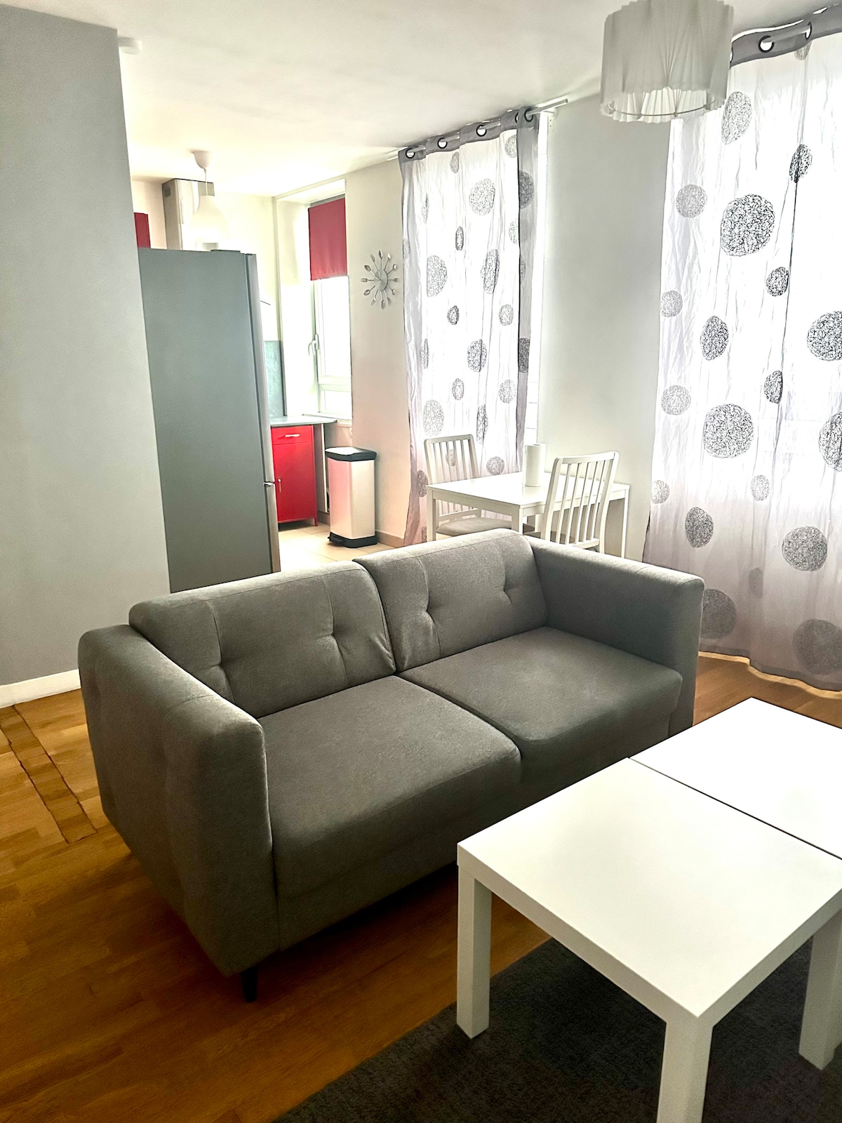 A comfortable gray sofa is positioned in the living area, complemented by a simple white coffee table. Bright curtains with circular patterns allow natural light to fill the space, which includes a small dining table in the background, enhancing the open layout.