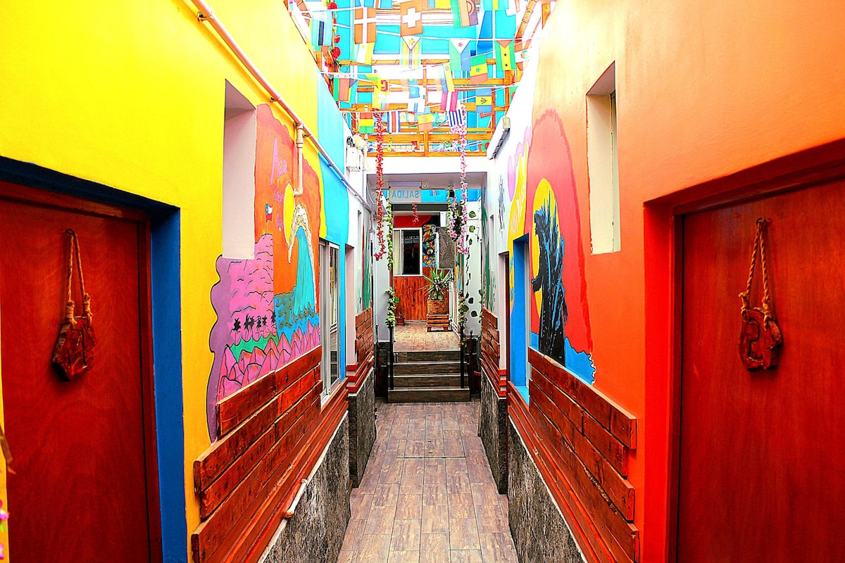 A vibrant corridor showcases walls painted in bright colors with artistic murals. Wooden doors lead to individual rooms, with decorative touches like hanging ornaments. Soft lighting illuminates the space, creating a lively atmosphere that invites guests to explore.
