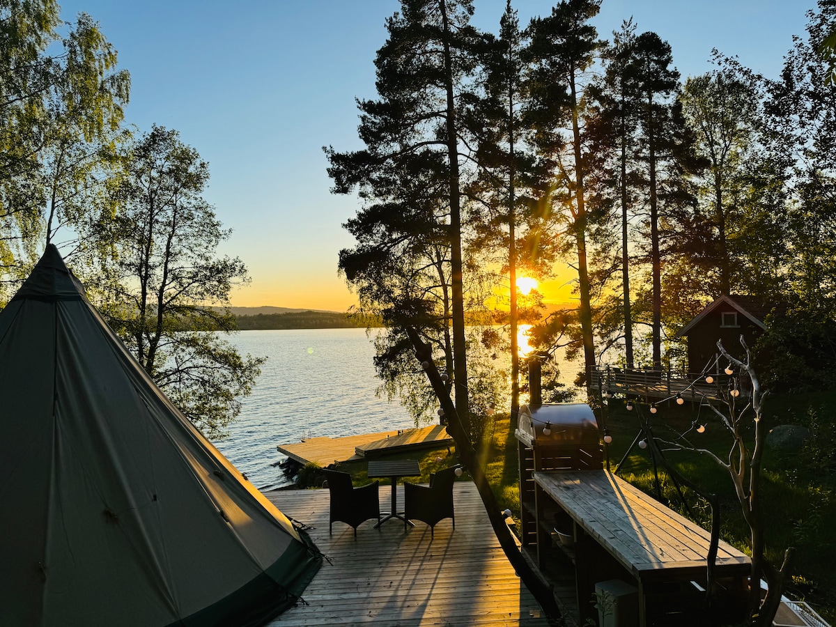 The image captures a scenic view of a lake during sunset, framed by tall trees. A tent is positioned on the left, while an outdoor dining area with a table and chairs is visible nearby. A wooden deck extends toward the water, enhancing the natural surroundings.