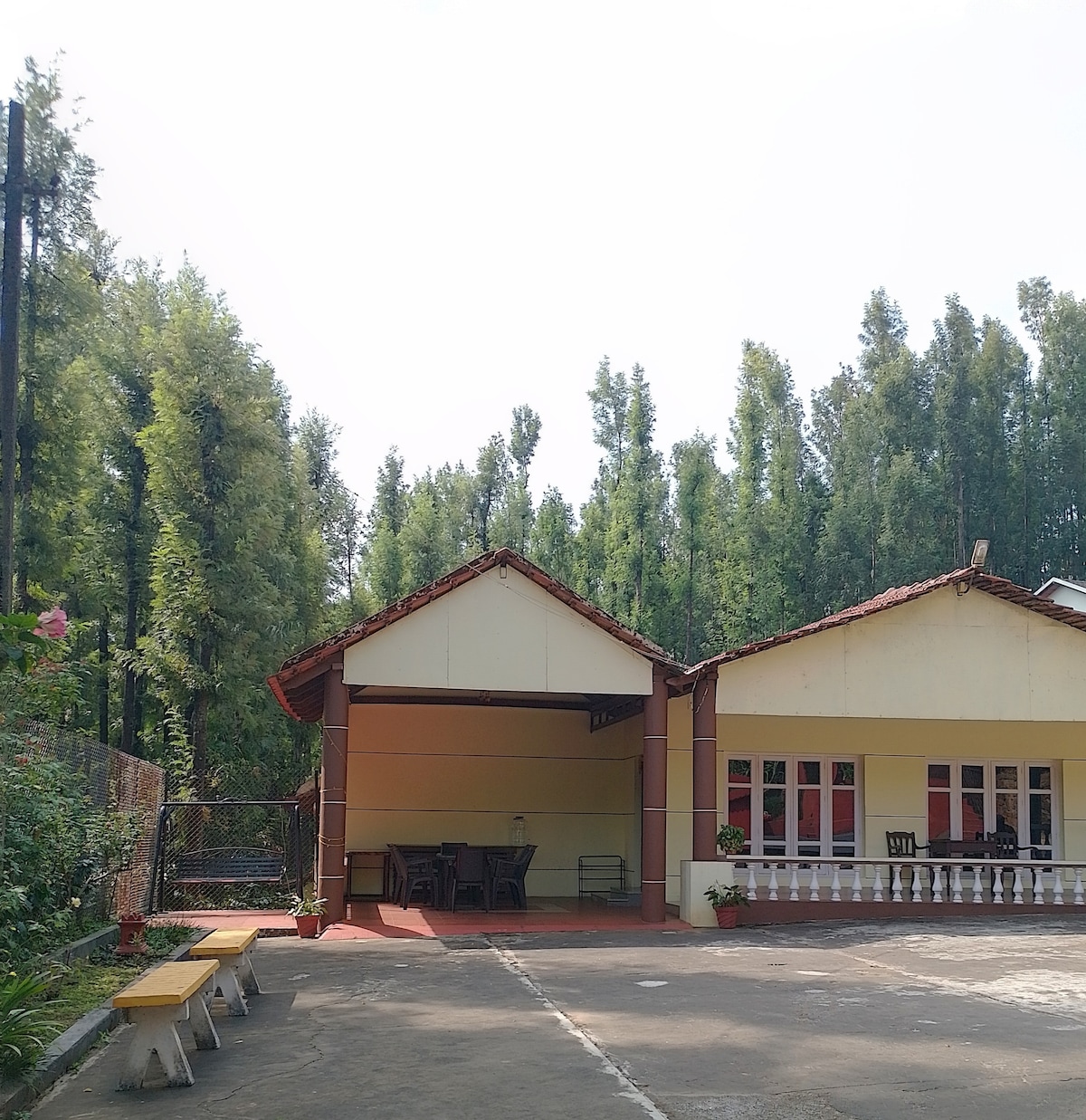 A welcoming independent house is set within a serene environment, framed by lush greenery. The exterior features a covered patio with seating and a distinct red roof. Multiple windows allow for natural light, enhancing the peaceful outdoor space surrounded by nature.