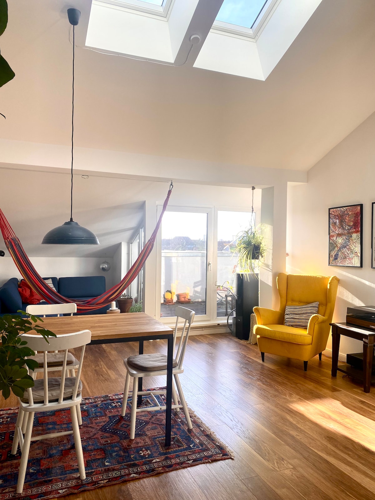 The dining area features a wooden table surrounded by several chairs, complemented by a vibrant area rug. A hammock is suspended above, inviting relaxation. Large glass doors open to the balcony, with natural light illuminating the space and highlighting the contrasting colors of the furniture.