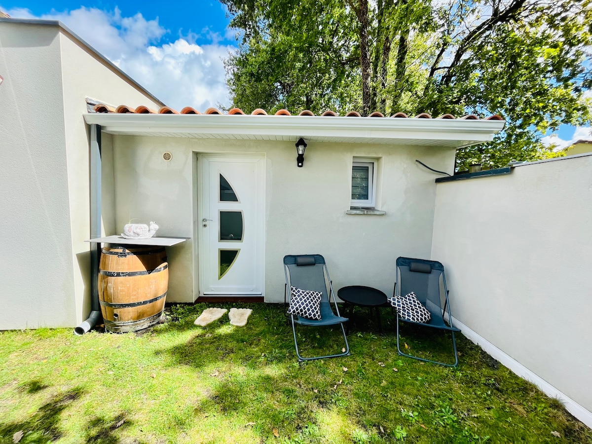 The exterior features a quaint entrance with a curved door and a small seating area. Two chairs with patterned cushions are positioned by a circular table, alongside a wooden barrel. The lawn is well-maintained, with sunlight filtering through the trees above.