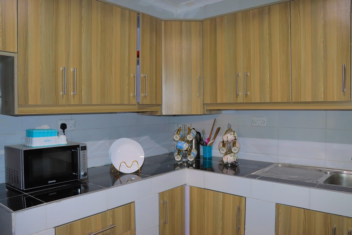 A modern kitchen features wooden cabinetry with sleek handles. A microwave sits on the countertop beside a decorative plate displayed on a stand. Colorful utensils are organized in a container, and a stainless steel sink is positioned near the corner, blending functionality and style.