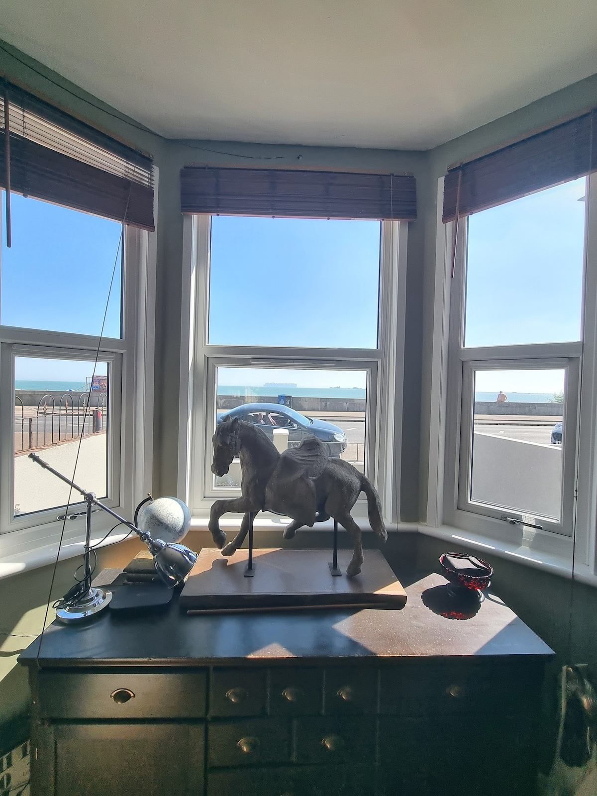 A corner window is filled with natural light, showcasing a decorative horse sculpture atop a dark wooden chest. A vintage radio and a small glass bowl are positioned nearby, with views of the beach and road visible through the windows.