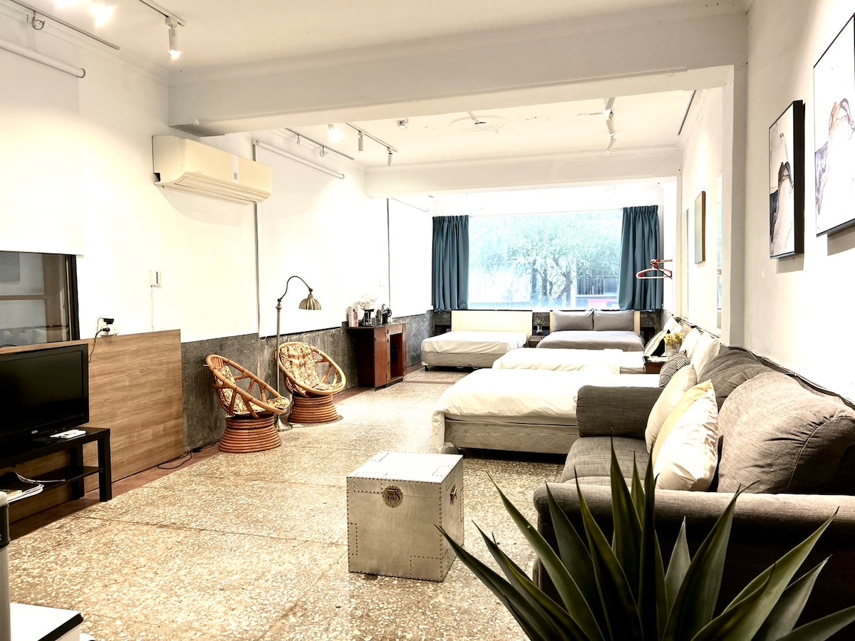 A spacious living area features a large window that allows natural light to illuminate the room. Comfortable seating options include a gray sofa and two rattan chairs. Two beds are positioned in the background, while a decorative metal table sits in the foreground.