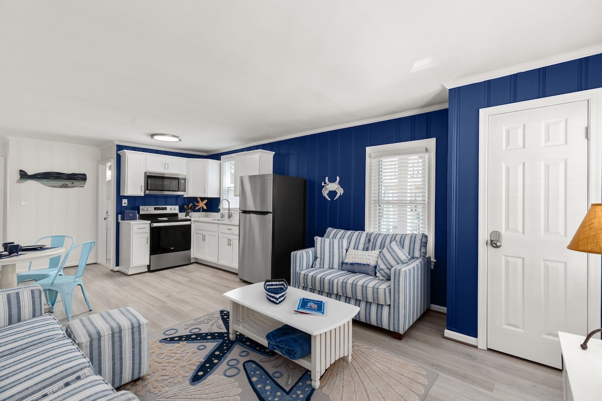 A spacious living area features blue accent walls, striped sofas, and a light wooden coffee table. A modern kitchen is equipped with stainless steel appliances and white cabinetry. Natural light fills the space through multiple windows, highlighting the coastal-inspired décor.