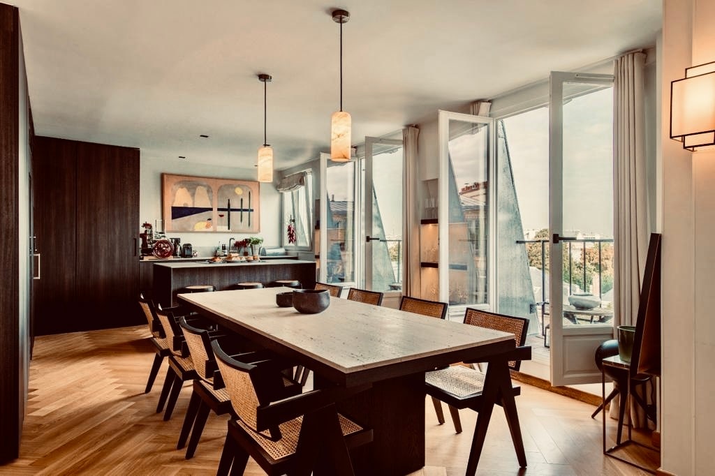 A spacious dining area is showcased with a long wooden table surrounded by eight woven chairs. Large glass doors open to a balcony, allowing natural light to flood the space. Modern pendant lights hang overhead, illuminating the elegant design.