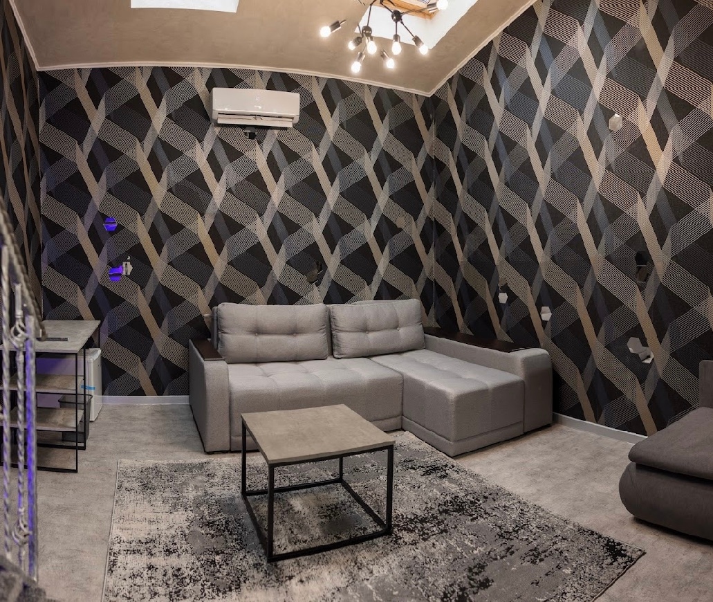A modern living area is presented, featuring a spacious sectional sofa arranged around a low coffee table. The patterned wallpaper creates an interesting backdrop, while an air conditioning unit is mounted on the ceiling. Soft lighting is provided by a distinctive chandelier.