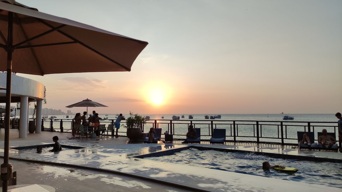 The pool area offers a clear view of the sunset over the sea, with lounge chairs under umbrellas and guests enjoying the space. The water reflects warm tones of the evening sky, while boats are visible in the distance.
