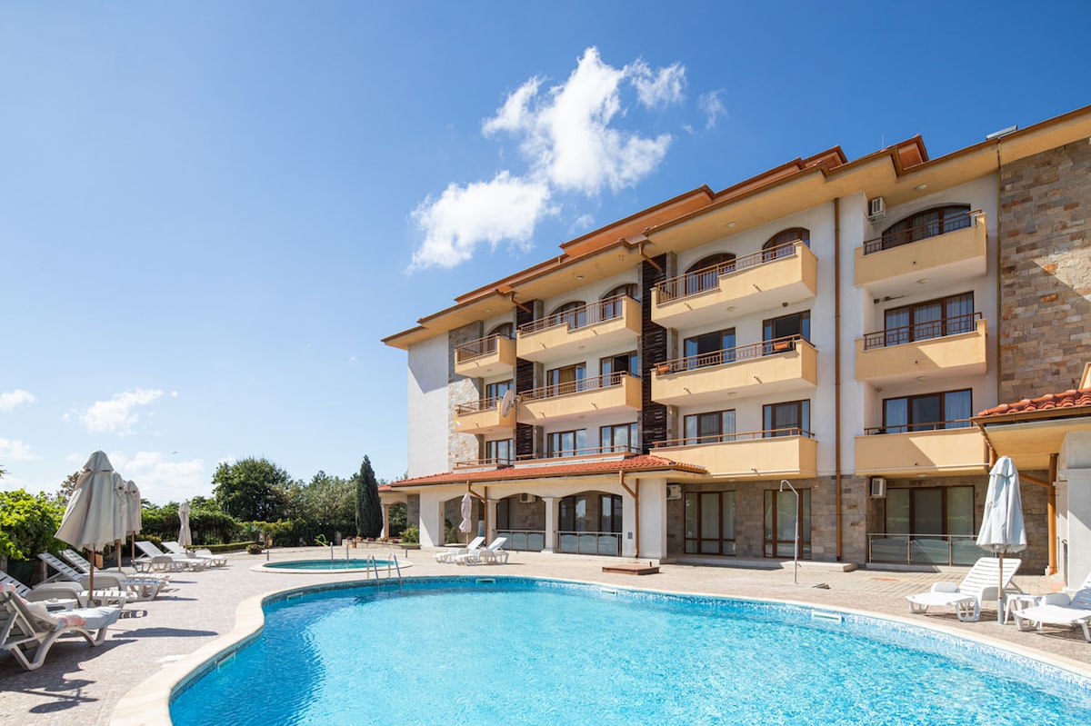 A well-maintained pool area is surrounded by lounge chairs and sun umbrellas, with a spacious multi-story building in the background. Lush greenery adds to the serene environment, while clear blue skies complete the picturesque setting.
