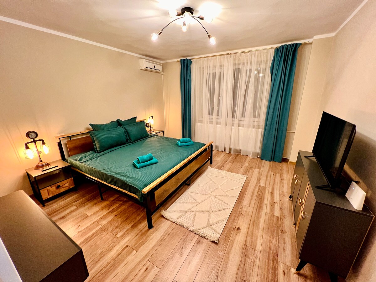 A well-lit bedroom features a spacious bed with teal bedding and pillows, complemented by two bedside lamps. A television is positioned on a black cabinet. Light drapes frame the large window, and a soft area rug lies on the wooden floor.