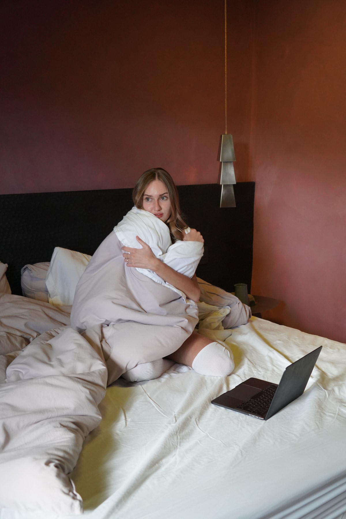 A spacious bed is covered with soft, neutral-colored bedding, creating a relaxed atmosphere. A laptop is positioned on the bedside, suggesting a comfortable space for work or leisure. The walls are painted in warm tones, contributing to a cozy environment.