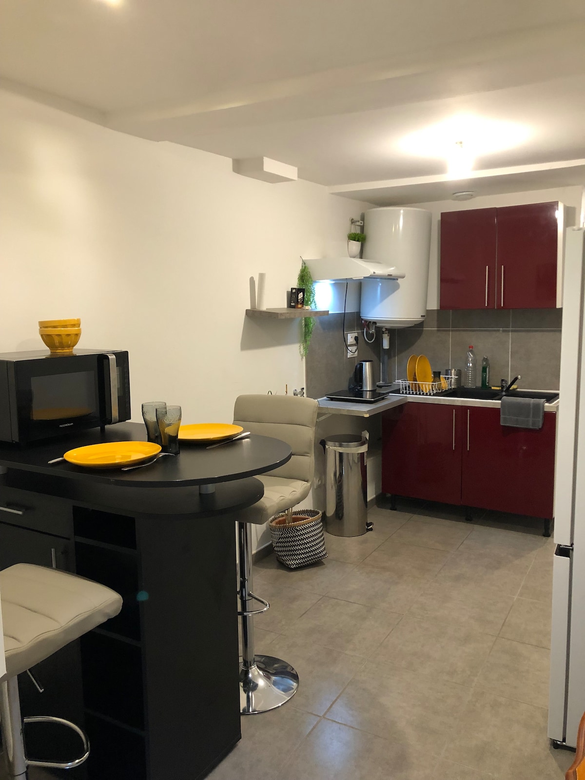 A compact kitchenette is featured with red cabinetry and modern appliances. A microwave and coffee maker are visible, accompanied by a sink and stovetop. A small dining area is set with two bar stools and yellow plates. The space is well-lit, showcasing ample storage options.