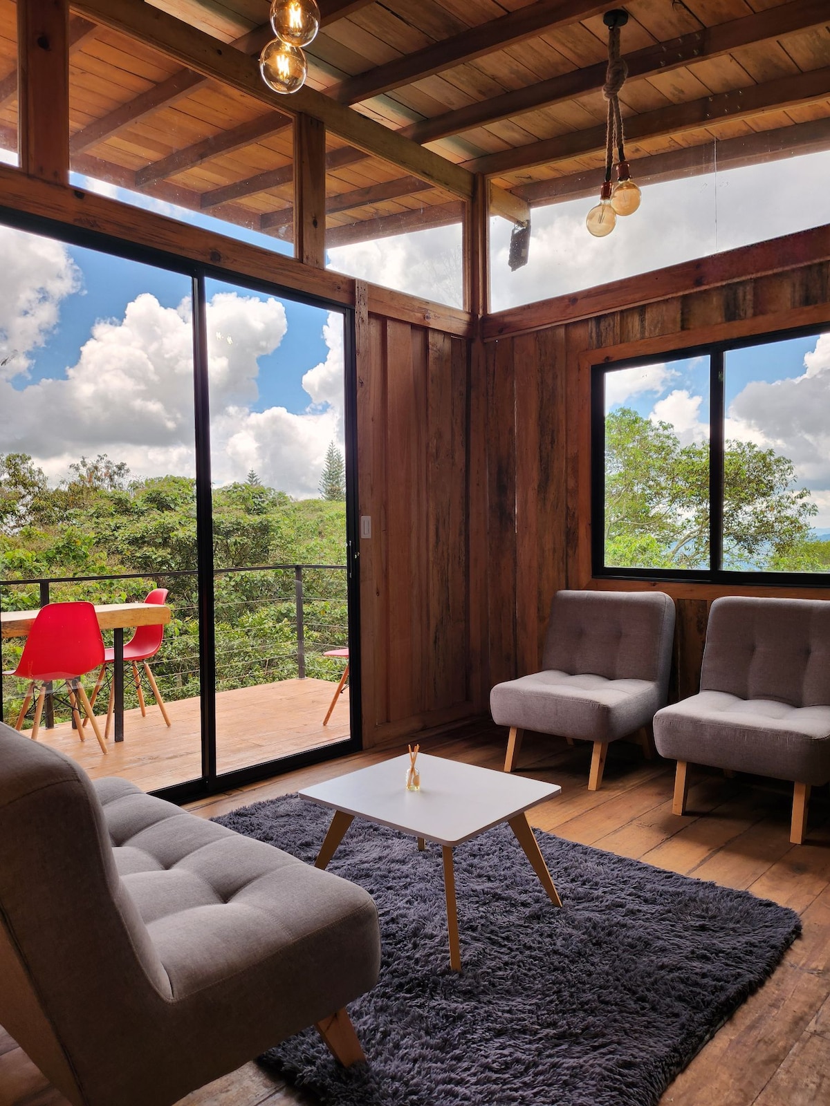 A cozy seating area is present with two gray armchairs facing a small white coffee table. Large glass doors lead to a deck with bright red chairs, offering views of lush greenery and a partially cloudy sky.