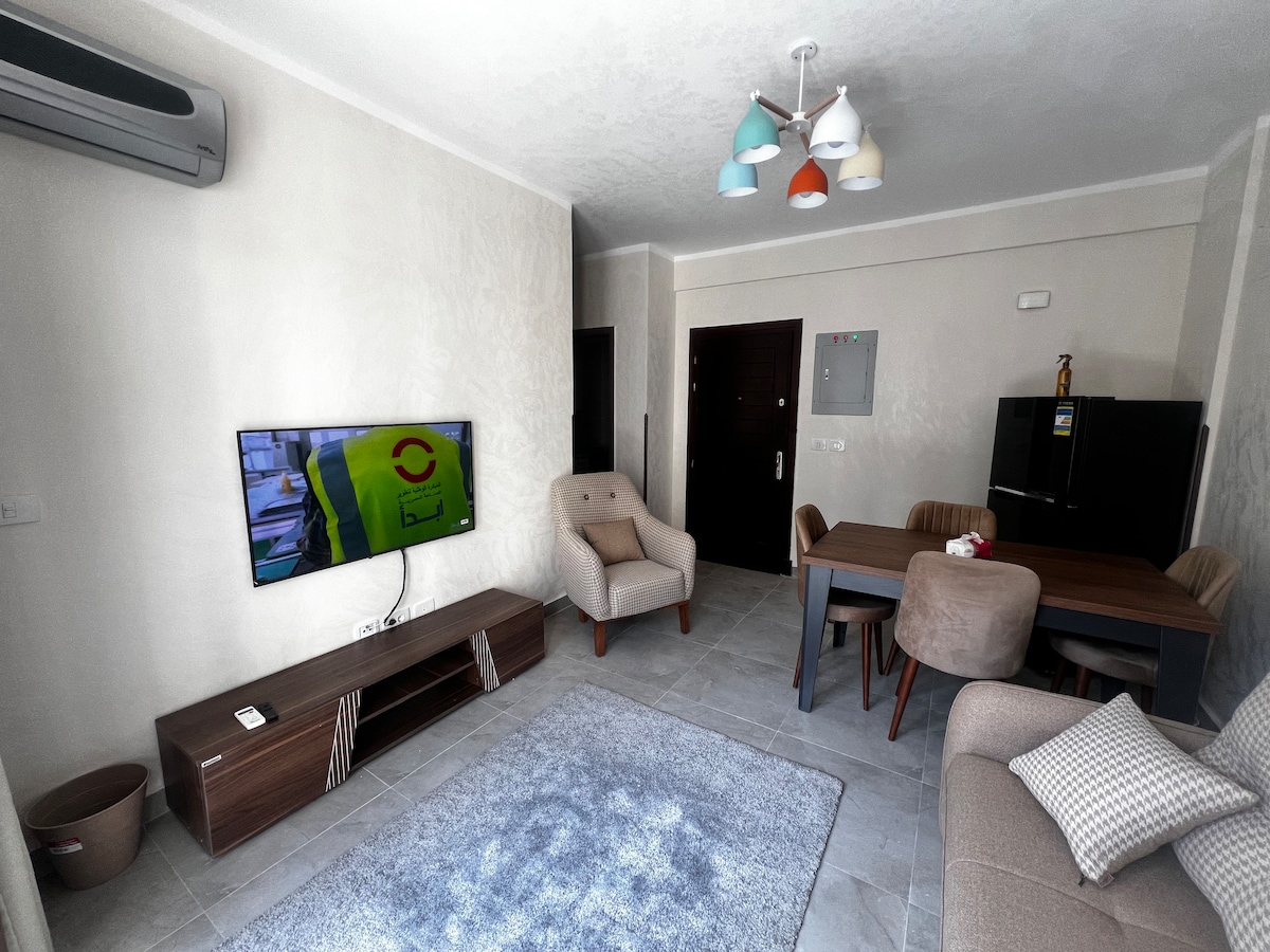 A modern living area features a light-colored sofa and two plush armchairs positioned near a large television. A dining table with upholstered chairs is visible, complemented by a grey rug underfoot. Brightly colored light fixtures hang from the ceiling, adding a touch of warmth.