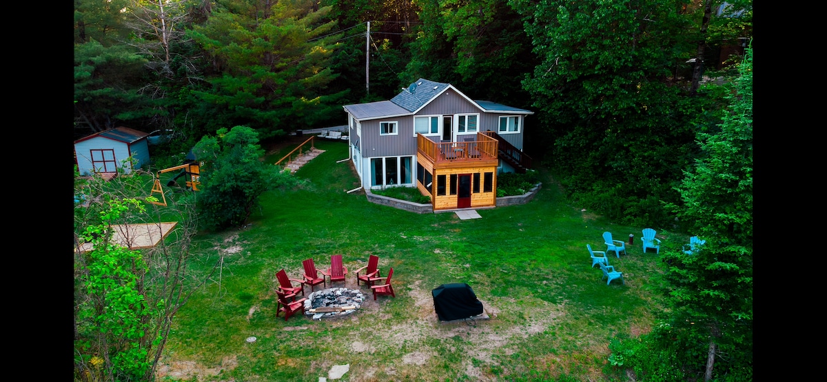 An aerial view of a two-story cottage surrounded by greenery. The outdoor area features a fire pit with six Muskoka chairs arranged around it, and a deck extending from the cottage. A large grassy lot with various seating areas is visible.