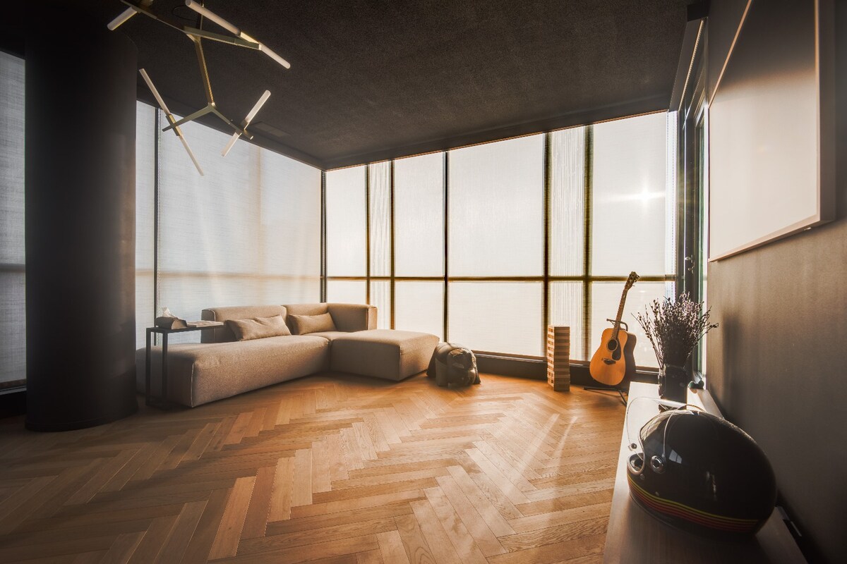 A spacious living area features large floor-to-ceiling windows, allowing natural light to illuminate the modern furnishings. A comfortable sectional sofa is positioned beside a decorative guitar and a sleek media console, while warm wood flooring adds an inviting touch.