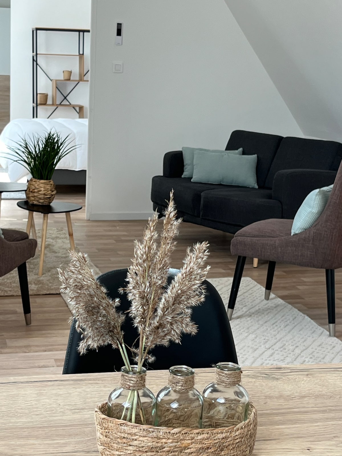 A stylish living area is visible, featuring a dark gray couch and a small round table. A woven basket holds three glass bottles with dried grasses. The background includes a glimpse of a bed and soft furnishings, accompanied by natural light filtering through the space.