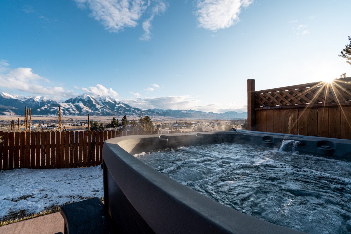 A hot tub is positioned on a patio with a frosty landscape visible in the background, showcasing distant mountains under a clear blue sky. The sunlight is seen streaming through the wooden fence, creating a serene outdoor atmosphere.