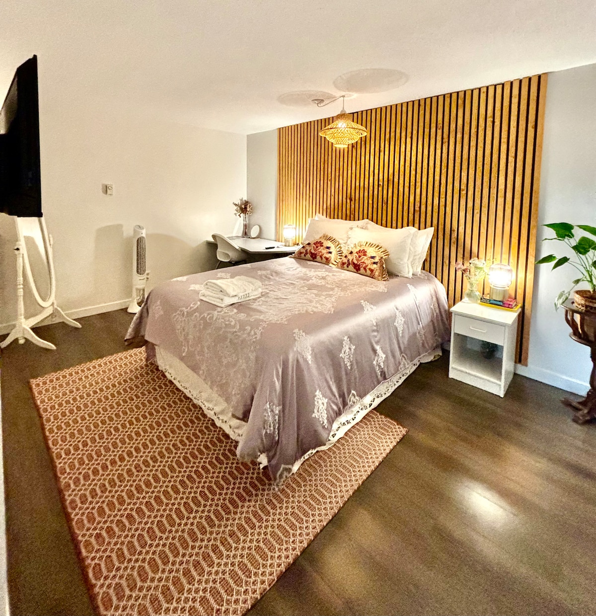 A well-appointed bedroom features a spacious bed with a light-colored quilt and decorative pillows. A wooden accent wall adds texture, while bedside lamps provide soft illumination. A small table holds amenities, and a decorative plant adds a touch of greenery. The room's warm tones create a relaxing atmosphere.