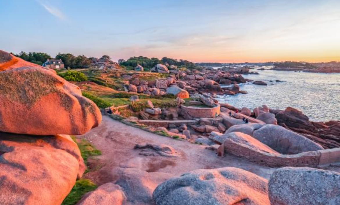 A rocky coastal landscape features large, smooth pink stones arranged along a shoreline. The scene is illuminated by a warm sunset, casting soft colors across the water. A pathway meanders among the rocks, leading to a scenic view of the sea and distant land.