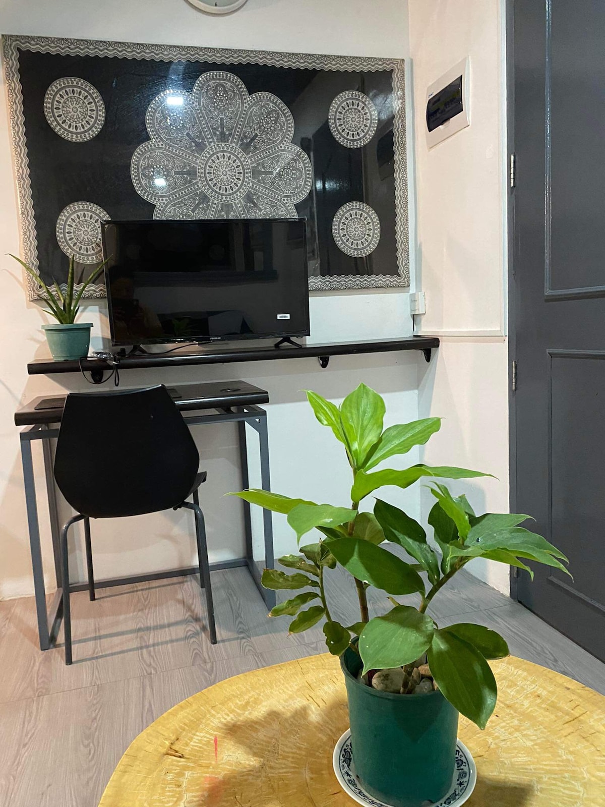 A workspace is featured with a wall-mounted television and a sleek desk paired with a chair. A decorative plant is placed on a round table, enhancing the space's greenery. A detailed black-and-white wall art is visible, providing a unique backdrop.