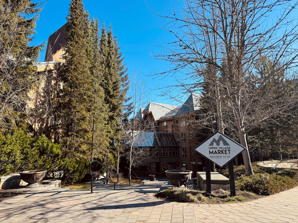 The entrance to the Upper Village Market is depicted, flanked by tall pine trees showing varying shades of green. A clear blue sky creates a tranquil backdrop, and a well-maintained pathway leads towards the market area, enhancing accessibility for visitors.