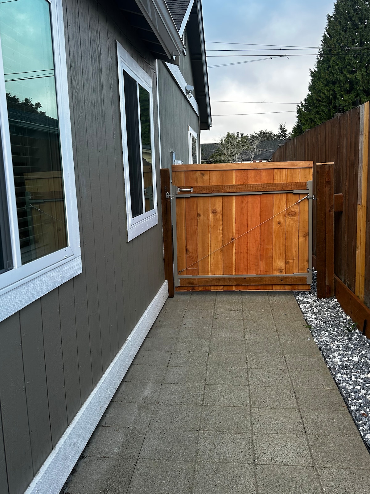 A narrow pathway is depicted, bordered by wooden fencing on either side. The ground is paved with square tiles, leading towards a wooden gate that stands slightly ajar. Two windows on the left side reflect natural light, enhancing the view of the outdoor area.