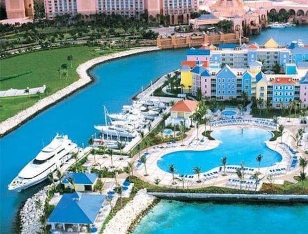 An aerial view captures the colorful facades of villas surrounding a large, curved pool. A marina filled with yachts is visible, along with lush green areas and smooth pathways. The vibrant landscape showcases the close proximity of water and greenery, creating a serene atmosphere.