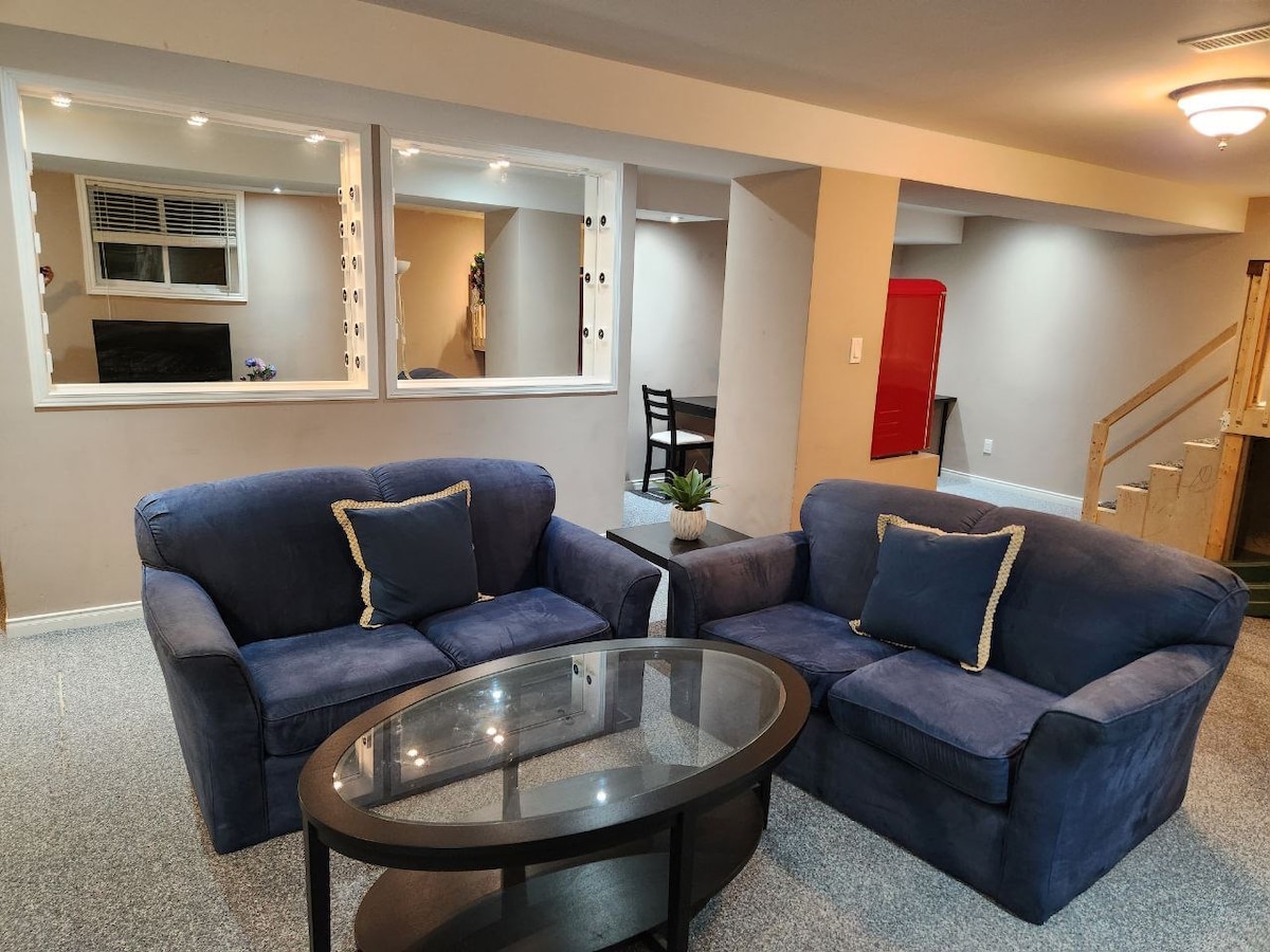 A cozy living area is furnished with two blue sofas and a circular glass coffee table. Natural light enters through a window and is reflected by a mirror above the seating area. A dining table is visible in the background, along with steps leading to additional spaces.