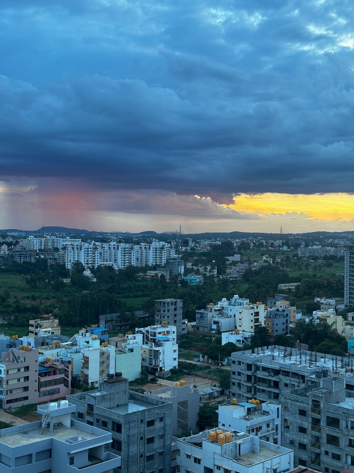 A panoramic view of the cityscape showcases a mix of residential buildings against a backdrop of rolling hills. The sky features dramatic clouds and a gradient of colors as the sun sets, creating a serene and picturesque atmosphere.