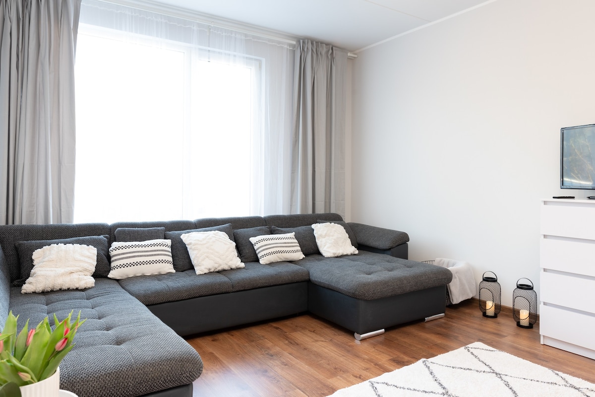 A spacious living area features a large sectional sofa adorned with decorative cushions. Sunlight filters through the expansive window, enhancing the light color of the walls and the warm wood floor. A small television on a white dresser and lanterns beside the sofa complete the setting.