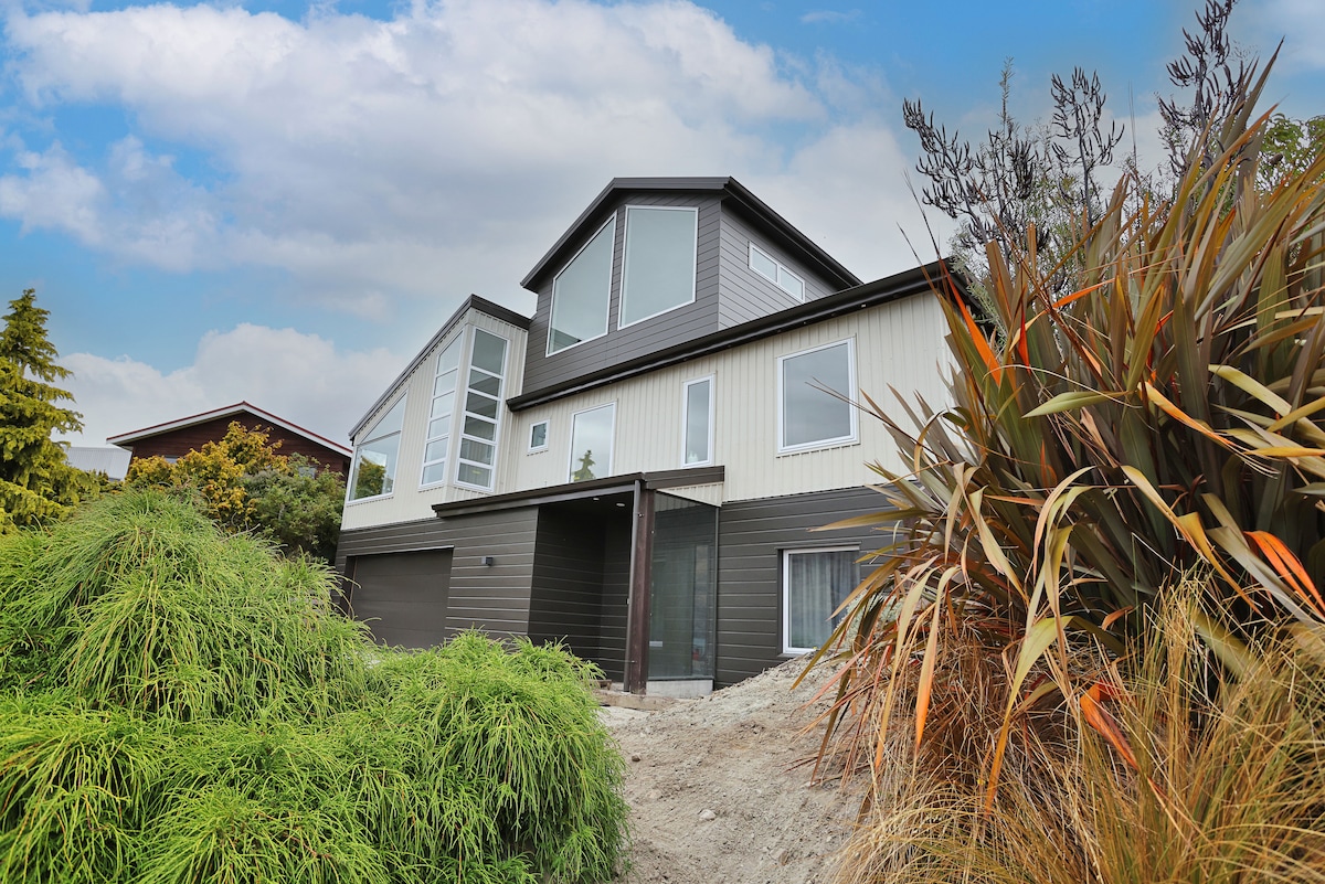 An architecturally designed home is presented, showcasing a combination of sleek lines and large windows. Surrounding foliage includes various green and orange plants, enhancing the natural setting. The exterior features a mix of light and dark cladding, set against a backdrop of a cloudy sky.