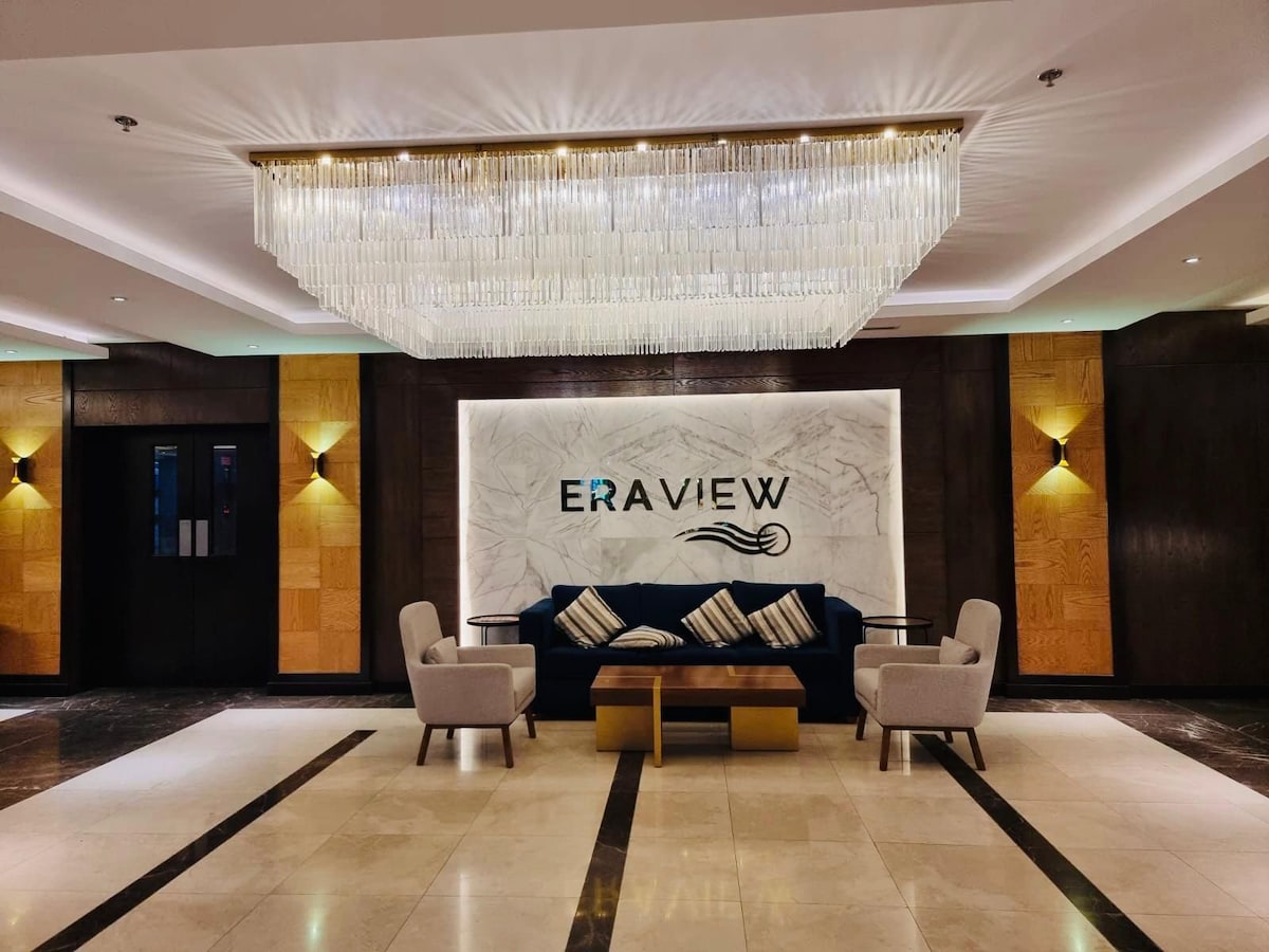 A modern lobby features a chic seating area with two armchairs and a coffee table. A large, elegant chandelier hangs from the ceiling, illuminating the refined decor of wood and marble. The prominent 'ERAVIEW' logo is displayed against a backlit marble wall.