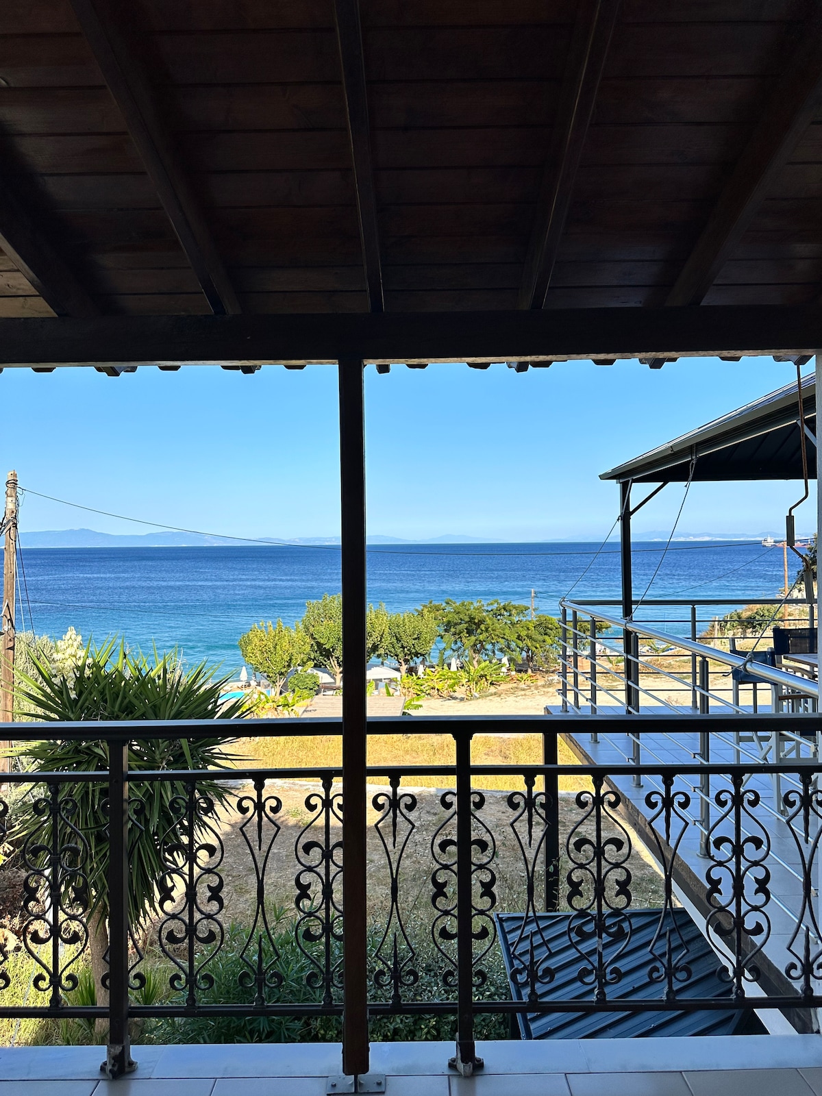A balcony view showcases a serene landscape of the sea and greenery. The clear blue water extends into the horizon, framed by a railing with delicate patterns. Sunlight casts natural light onto the outdoor space, creating a bright and open atmosphere.
