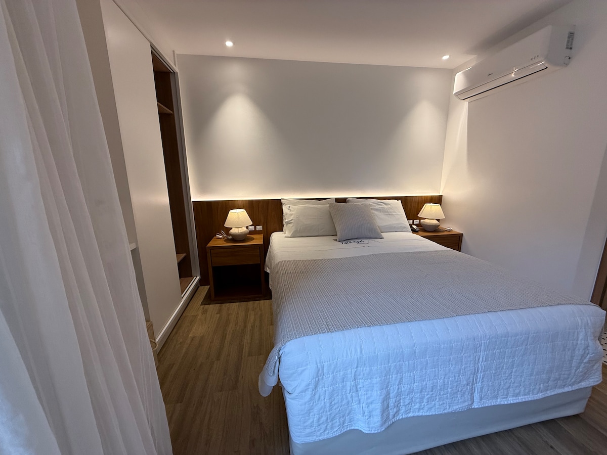 A well-lit bedroom features a spacious bed with a neatly arranged white bedspread. Two bedside tables with lamps add to the serene ambiance. A closet is present on one side, enhancing the room's minimalist design.