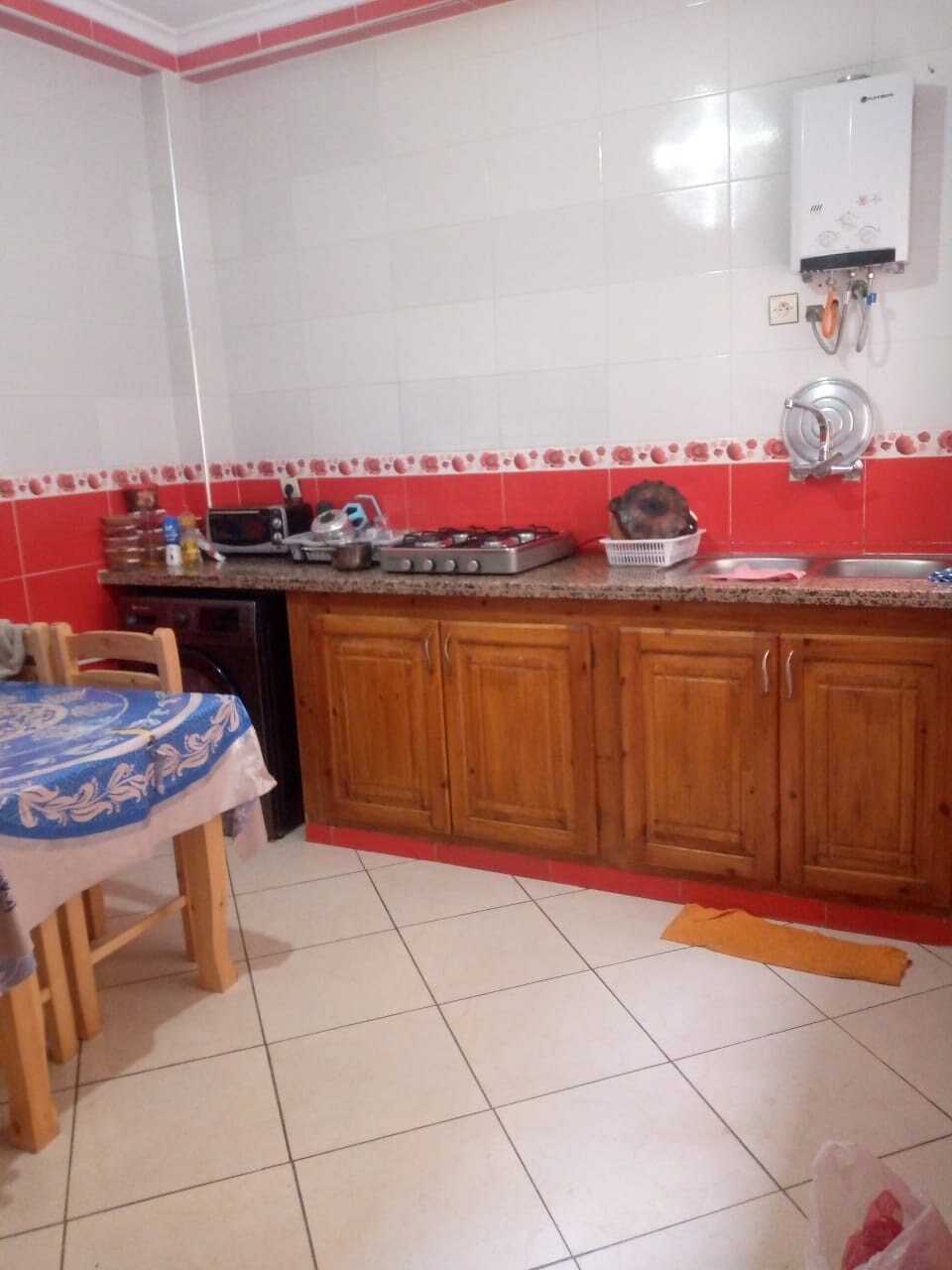 A functional kitchen space features wooden cabinets and a granite countertop. Appliances are arranged along the wall, with a gas stove visible. A small dining table with a patterned tablecloth is present, along with a bright orange mat on the tiled floor.