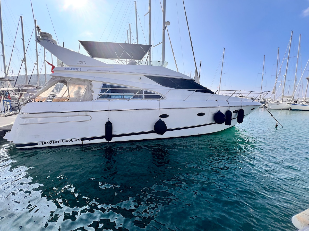 A sleek motor yacht is docked in a marina, featuring multiple sails and spacious deck areas. The hull is painted white, and the water surrounding the boat displays a clear blue hue, reflecting the sunlight.