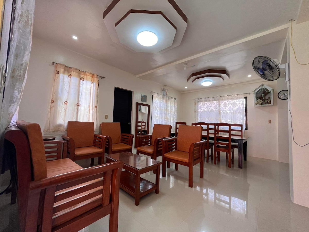 A spacious common area is showcased, featuring several wooden chairs and a coffee table. Natural light filters through sheer curtains, highlighting the clean, tiled floor. Ceiling fans are installed for comfort, creating a functional space for relaxation and gathering.