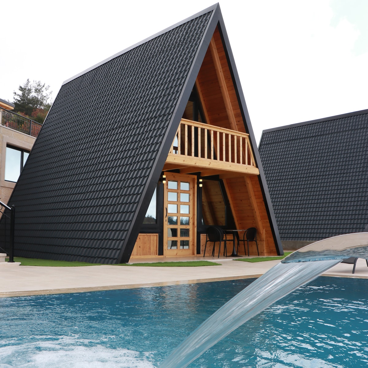 A striking triangular bungalow features a textured black exterior and a welcoming wood facade. A balcony with two chairs overlooks a serene swimming pool, while smooth water flows gently from a spout nearby, creating a tranquil environment.