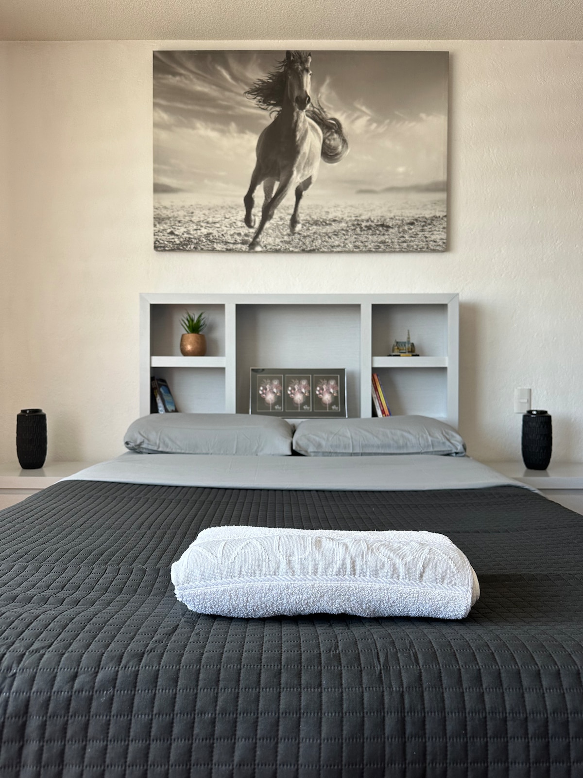 A spacious sleeping area is showcased, featuring a large bed with gray bedding. Above the bed, a monochrome artwork of a horse running adorns the wall. Bedside tables are positioned on either side, with decorative items placed on the shelves and a rolled towel resting on the bed.