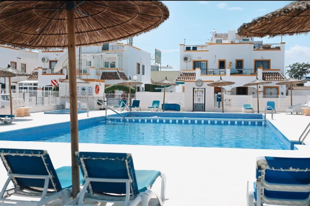 A refreshing swimming pool is surrounded by sun loungers and shaded areas under straw umbrellas. The pool's crystal-clear water reflects the blue sky, while nearby white buildings provide a backdrop that suggests a peaceful atmosphere.