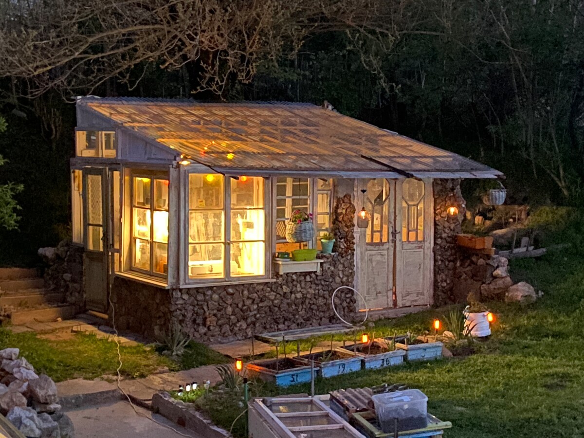 A charming greenhouse structure is illuminated at dusk, showcasing a combination of glass windows and natural stone. Two wooden doors, adorned with circular accents, lead inside. Soft lighting highlights the surrounding garden area, enhancing the serene atmosphere of the getaway.