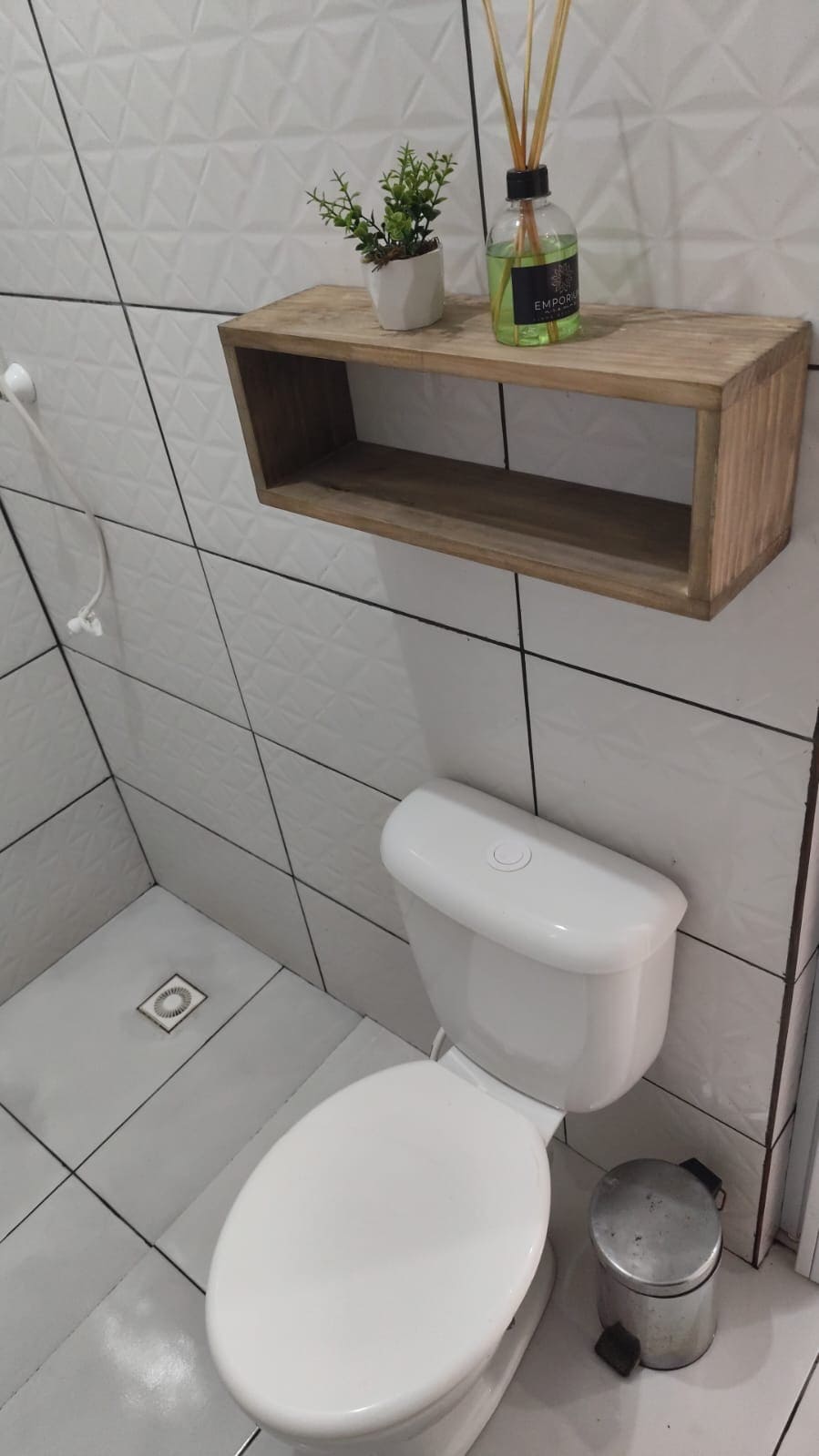 The bathroom features modern white tiles covering the walls and floor. A functional toilet is positioned in the foreground, while a wooden shelf above holds a small potted plant and decorative diffuser. A waste bin is visible near the toilet.